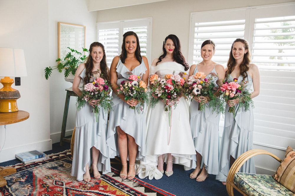 Bridesmaids in light gray dresses holding colorful floral bouquets pose together.