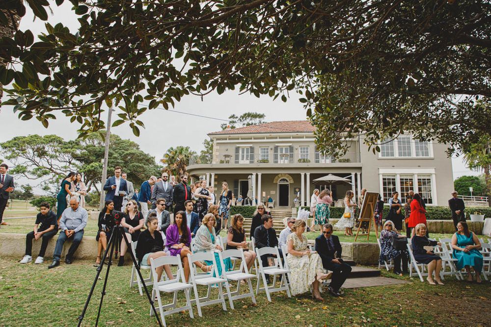 Outdoor wedding ceremony setup with white chairs under trees by the water.