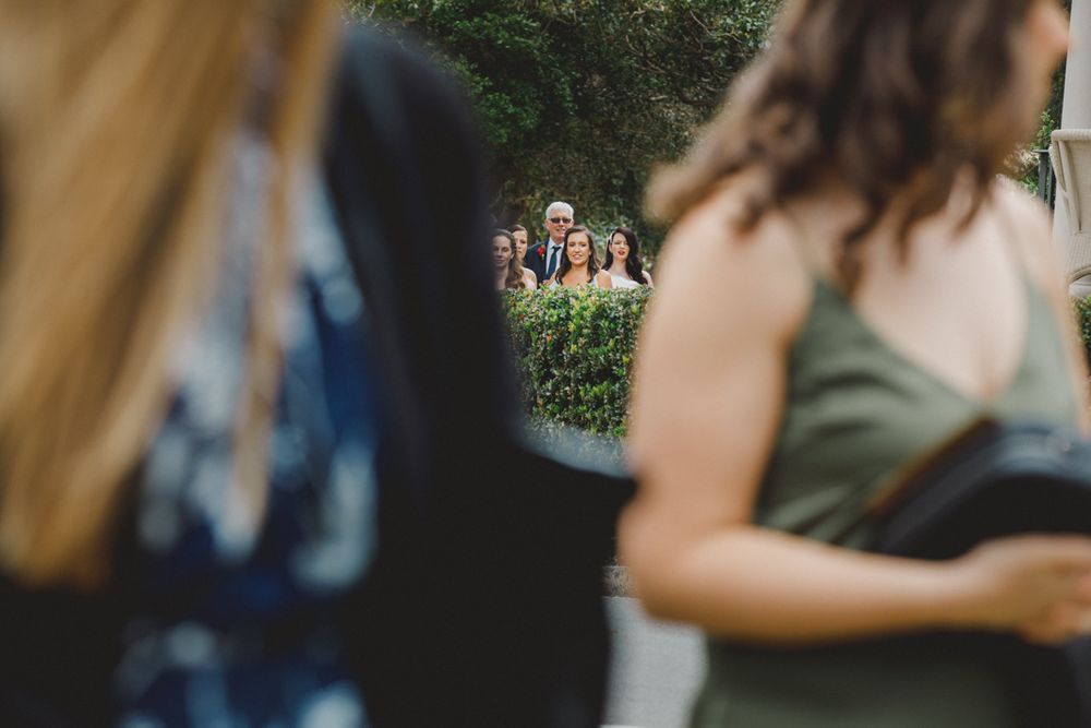 Guests can be seen in the background through a blurred foreground at an outdoor wedding ceremony.