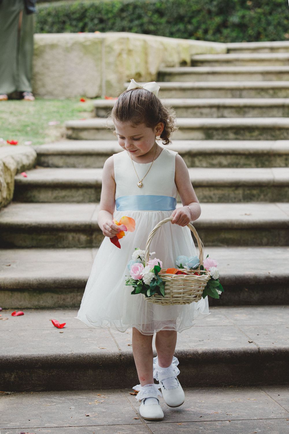 A flower girl in a white dress carries a basket while walking down stone steps.