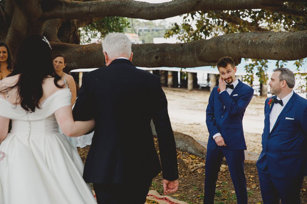 A wedding ceremony unfolds under a large tree branch in a garden setting.