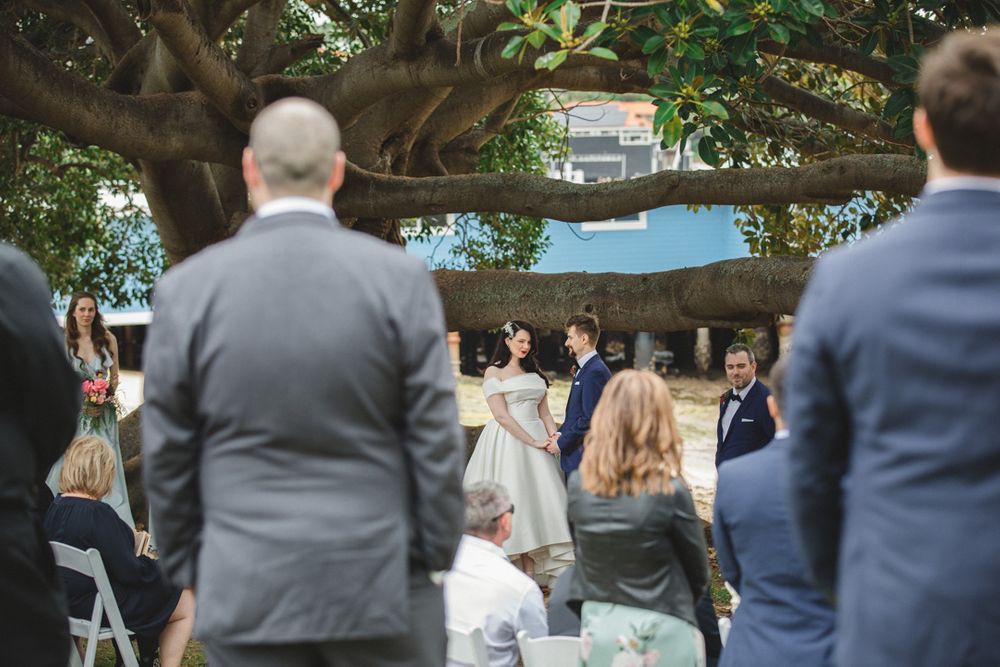 Guests watch an outdoor wedding ceremony beneath a sprawling tree.