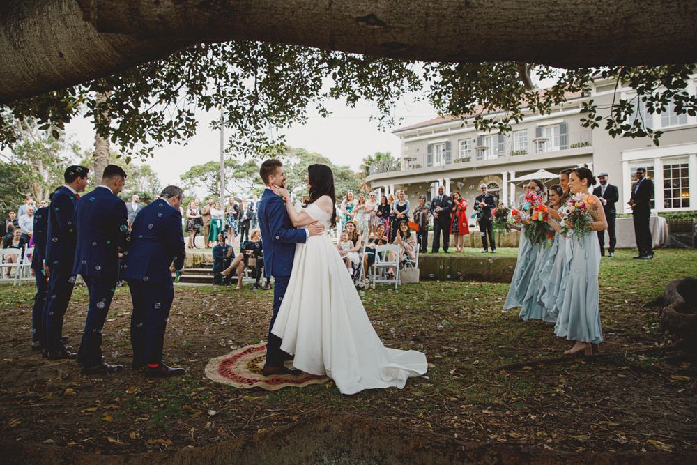 Wedding party gathers under trees for outdoor celebration with light blue and navy color scheme.