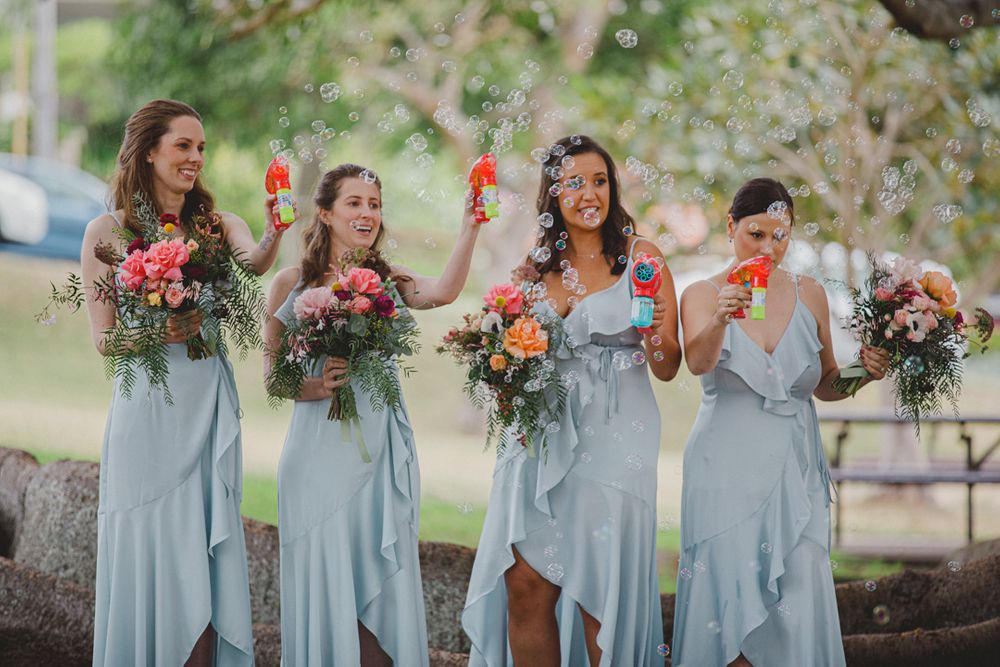 Bridesmaids in flowing light blue dresses hold colorful bouquets while celebrating outdoors.