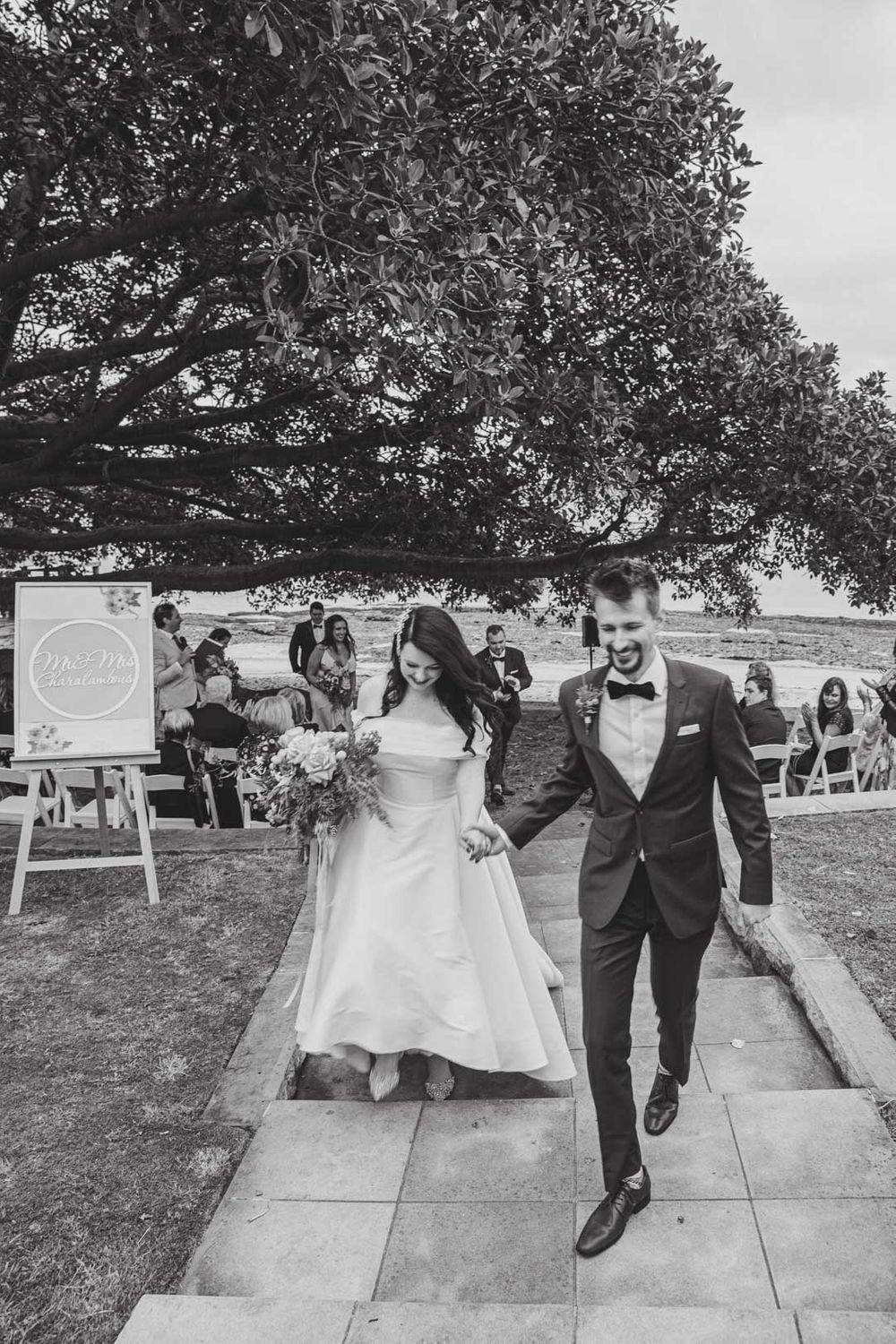 Black and white photo captures wedding party walking along outdoor pathway under large tree.