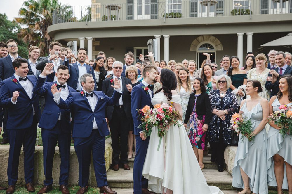 Large wedding party poses for group photo in front of elegant white building.