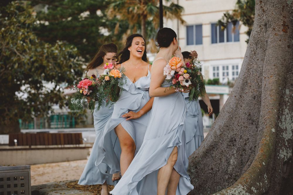 Wedding party in light blue dresses laughing together under a large tree in an outdoor setting.