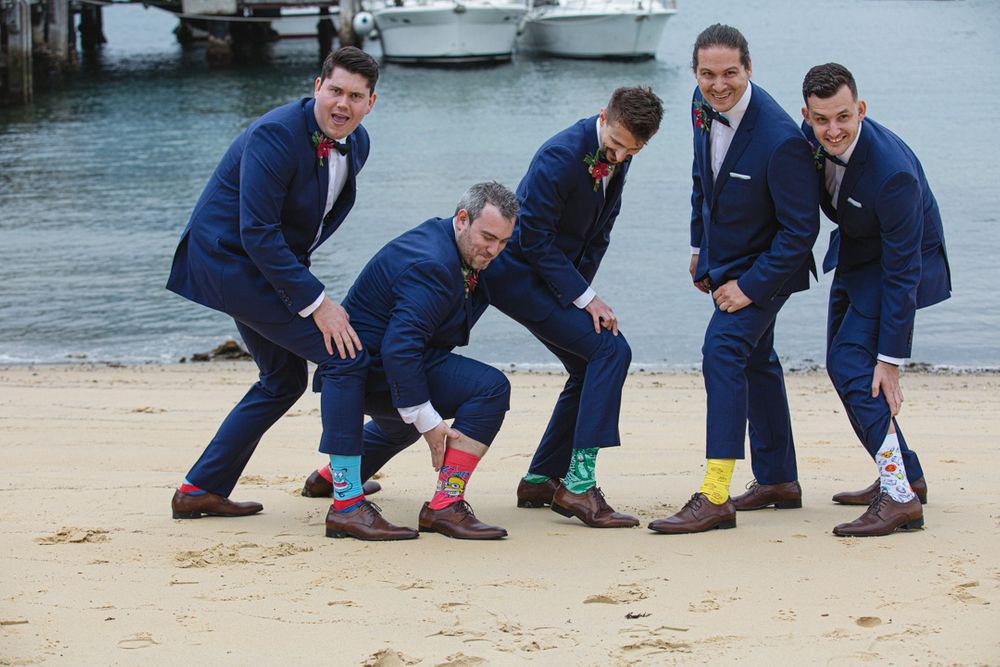 Groomsmen in navy suits showing off colorful socks while crouching on beach.