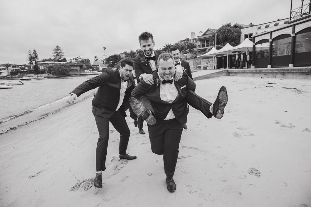 Wedding party performs synchronized jumping poses on sandy beach in black and white.