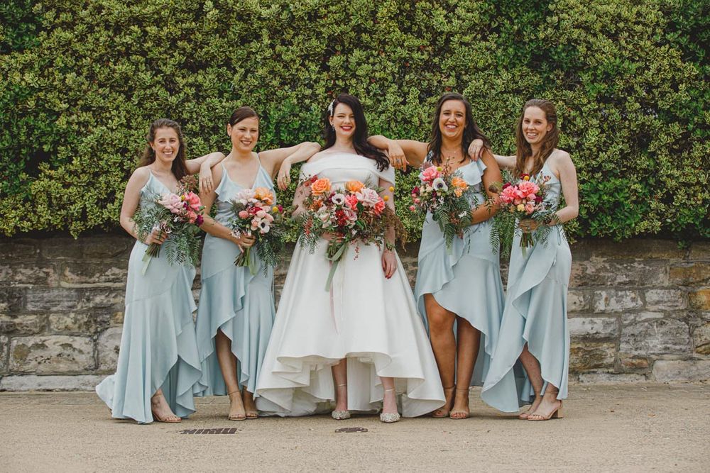 Bridesmaids in light blue dresses holding bouquets pose together against stone wall.