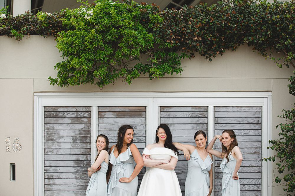 Wedding party posing against a white wooden wall with green foliage overhead.
