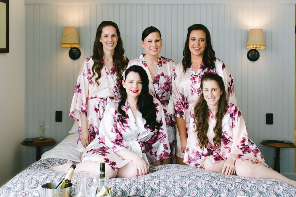Five women in matching pink floral robes pose together on a bed.