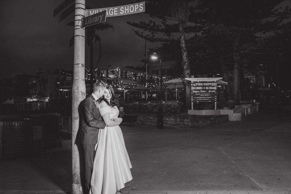 A romantic black and white nighttime scene showing a couple embracing under a street sign.