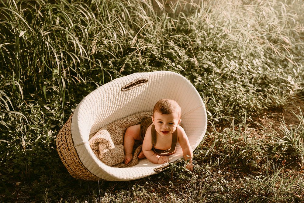 Baby playing in white woven Moses basket outdoors during golden hour.