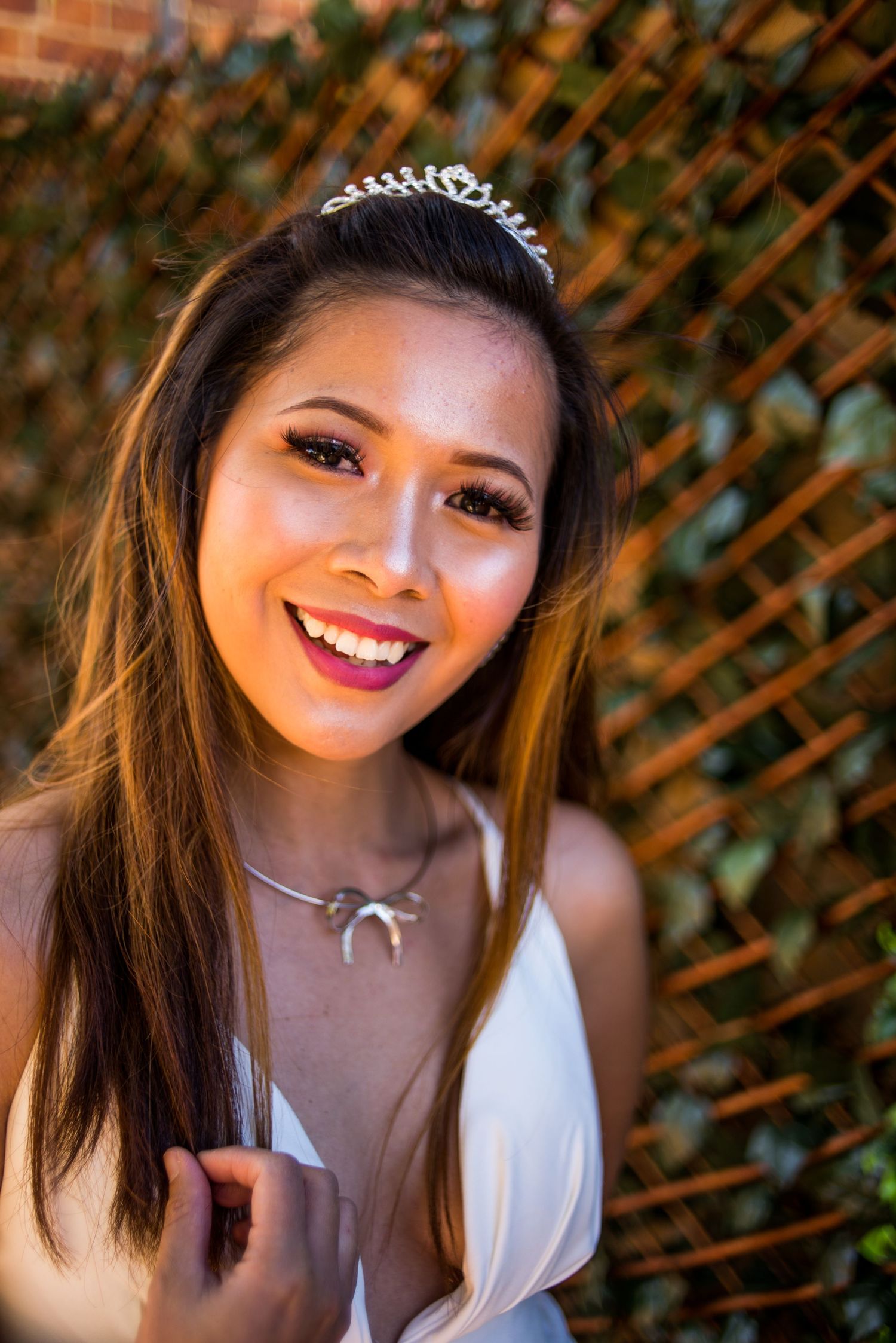 A radiant bride wearing a delicate tiara and white dress smiles in front of a latticed garden backdrop.