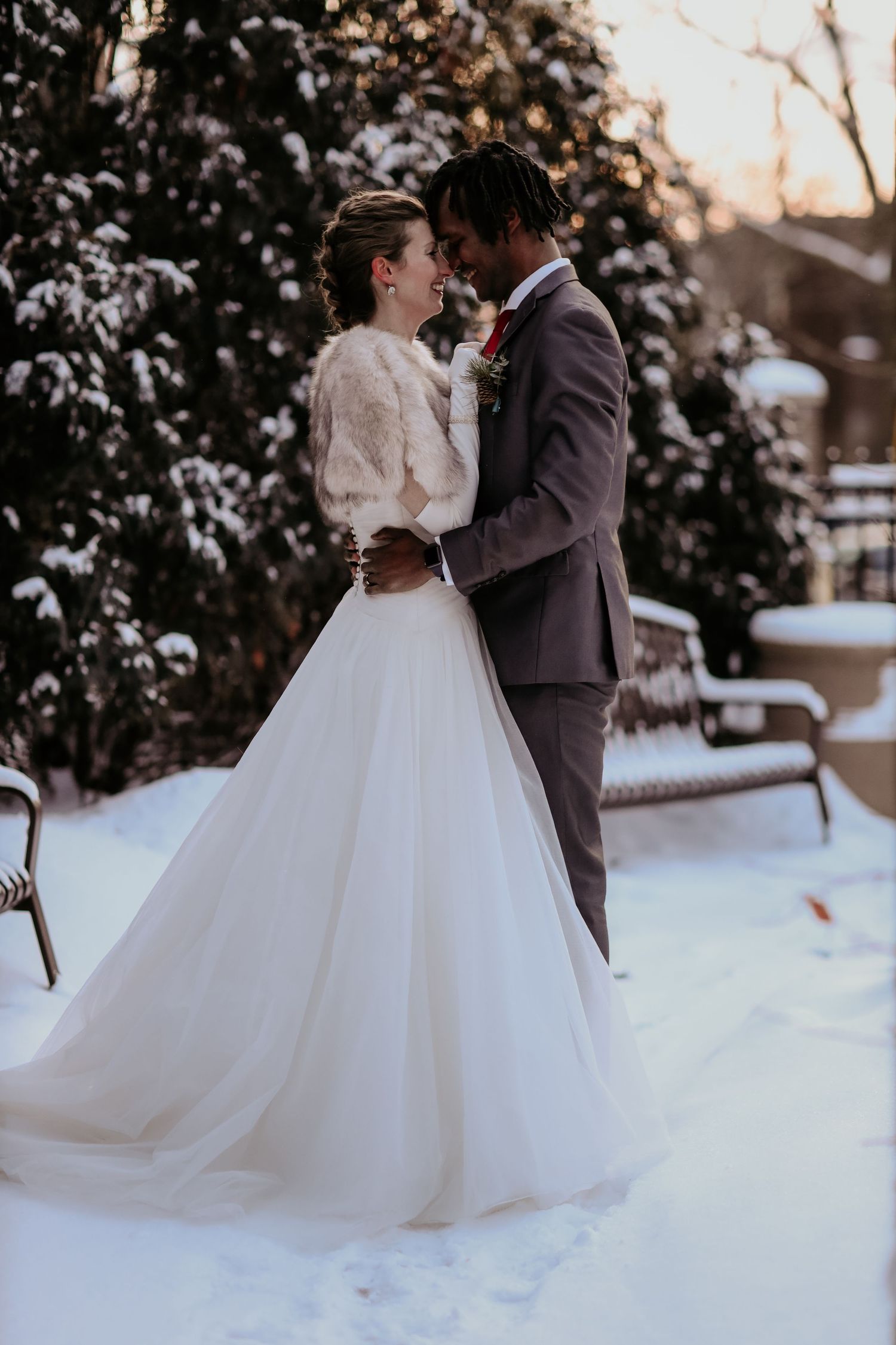 Winter wedding photo of couple embracing in snow with bride wearing long white dress and fur jacket.