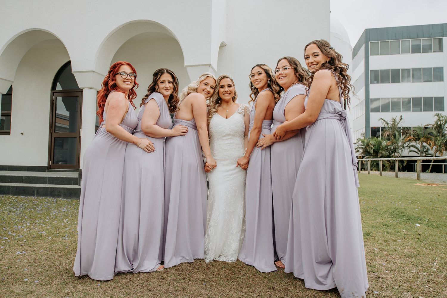 Bridal party wearing flowing lavender gowns poses on church lawn.