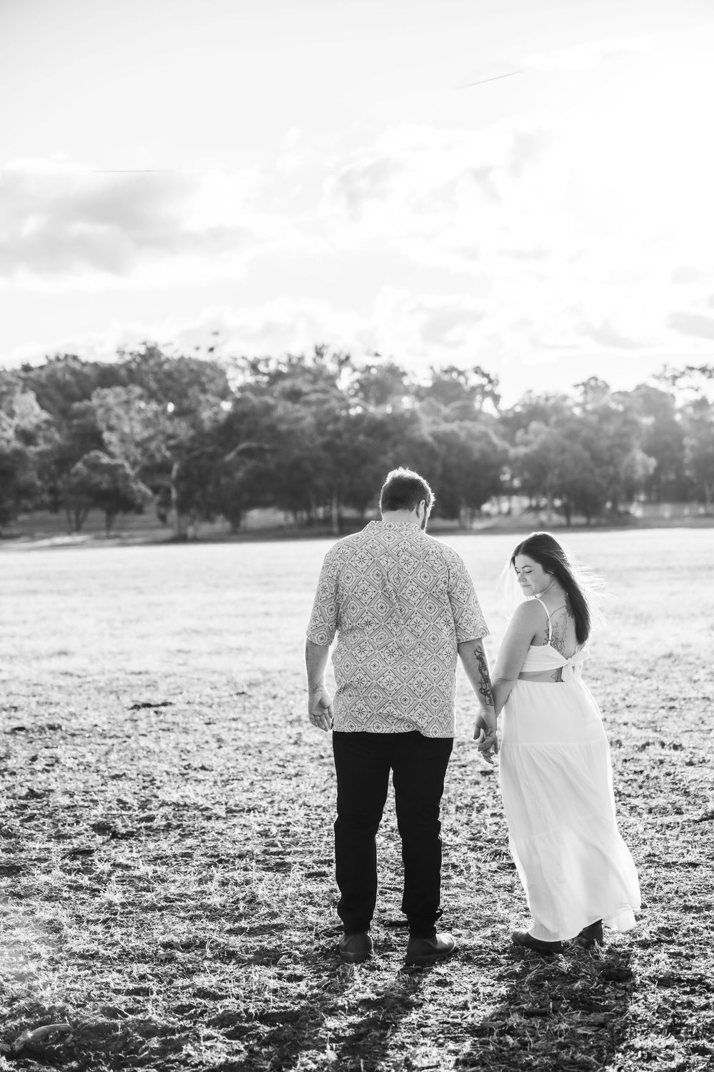 A couple walks hand in hand through a sunlit field with trees in the background.