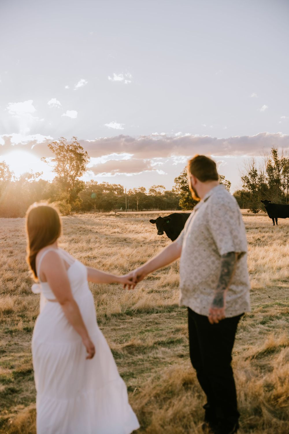 A couple holds hands while walking through a rural field at sunset with clouds in the sky.