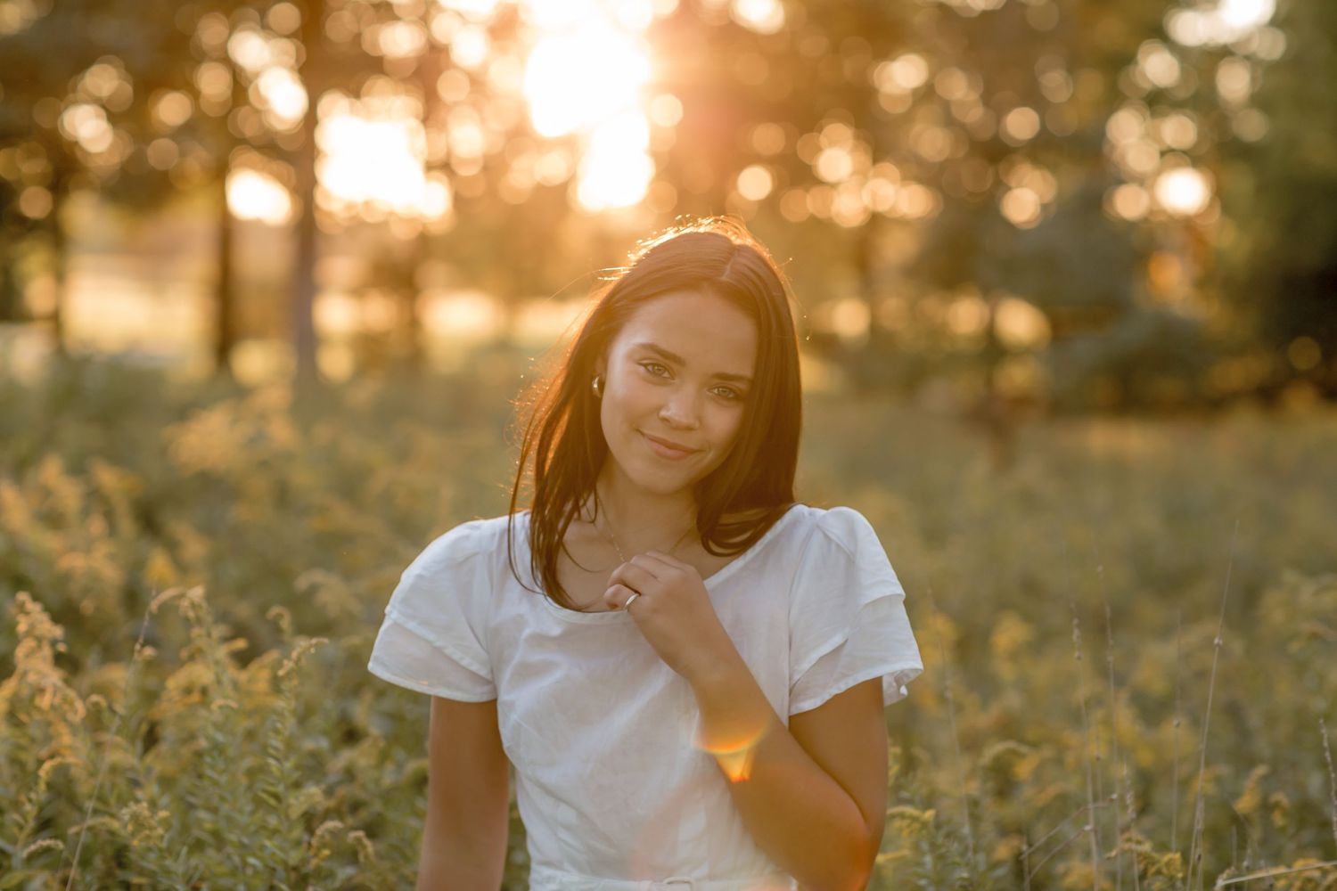 Young brunette woman in a white t-shirt standing in a golden field at sunset with warm backlight.