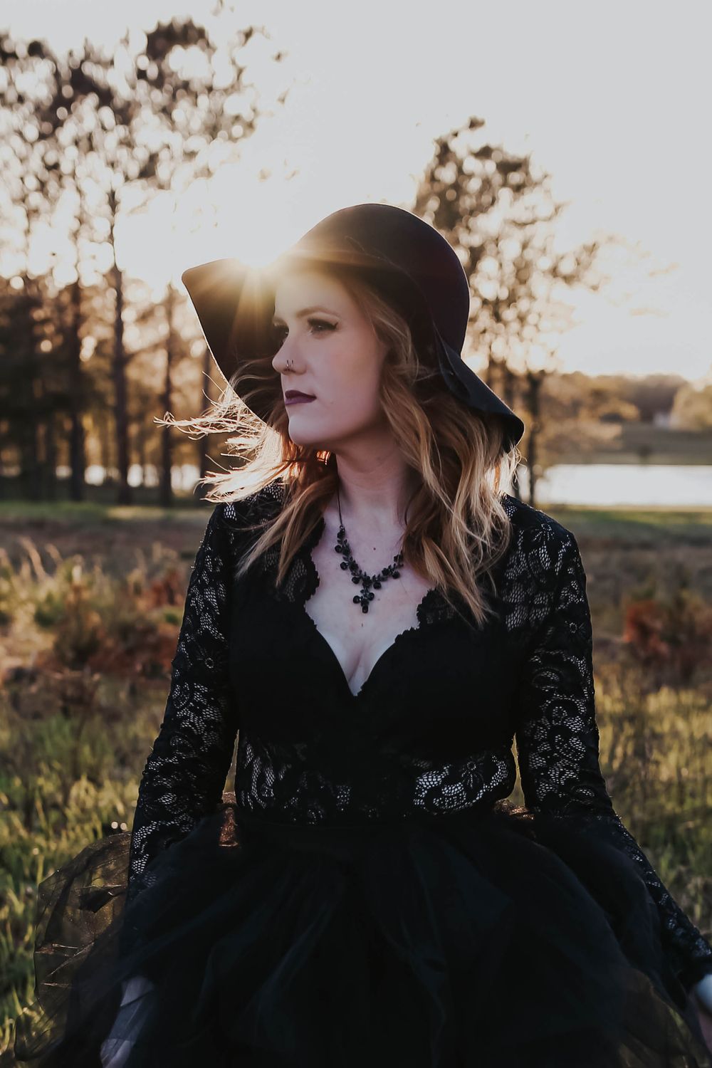 Ethereal portrait in black lace dress and hat with golden sunlight backdrop.