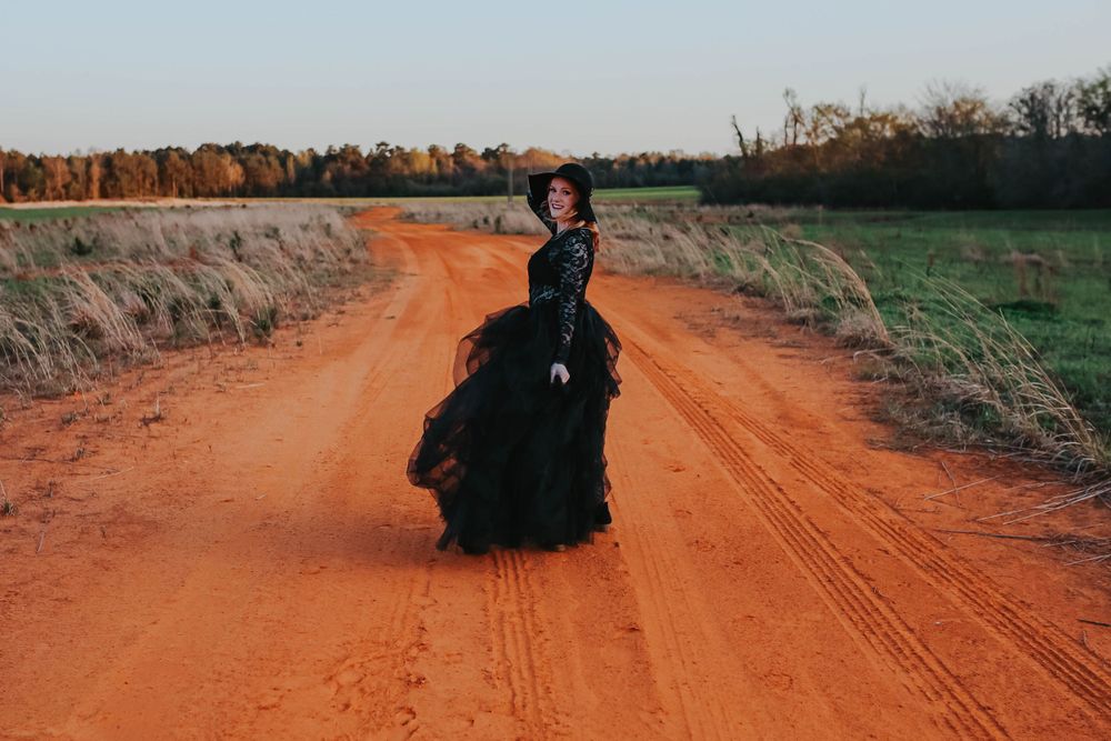 A figure in a dramatic black dress poses on a rustic orange dirt road surrounded by autumn fields.