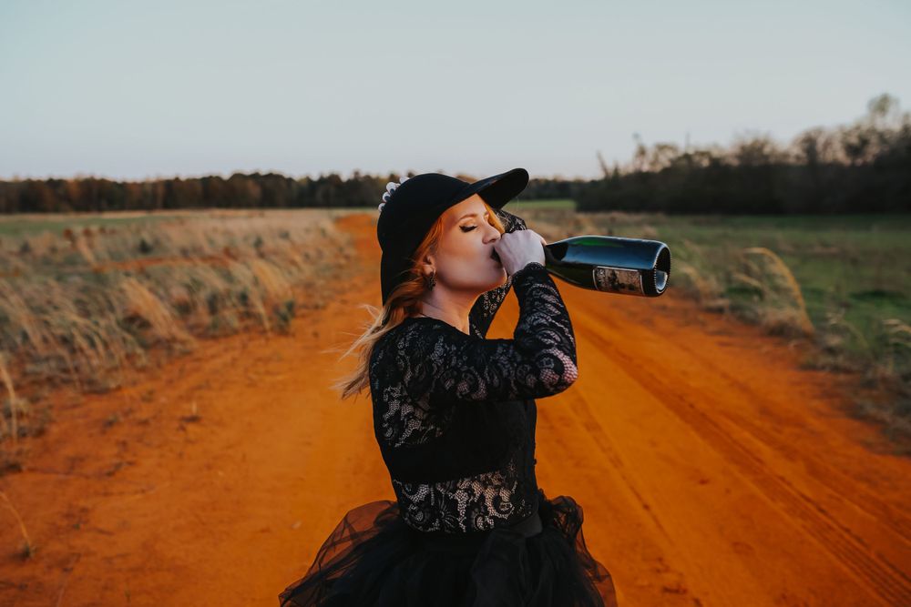 A person in black attire drinks from a bottle while standing on an orange dirt road at sunset.