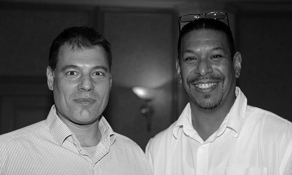 Black and white photo of two men posing together, one wearing a light shirt and one in a white shirt.