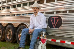 Cowboy in white hat sits with Australian Shepherd dog beside silver livestock trailer.