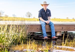 Young man in cowboy hat sits on wooden fence over marshy water in rural landscape.