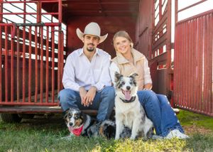 Couple sitting with two dogs in front of a red barn, man wearing cowboy hat, woman smiling, farm setting.