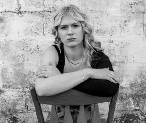 Black and white portrait of blonde woman sitting on chair against stone wall with arms crossed.