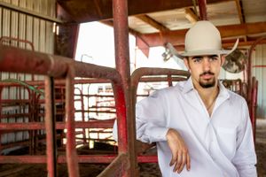 Young man in white shirt and cowboy hat leaning on rustic red metal gate in a barn setting.