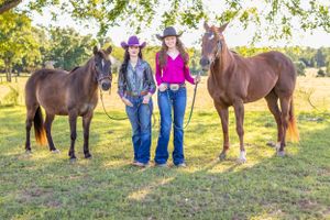 Two cowgirls in hats and jeans stand with brown horses in sunny green pasture.