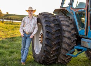 Young cowboy in straw hat leans against large tractor tire in green field at sunset.