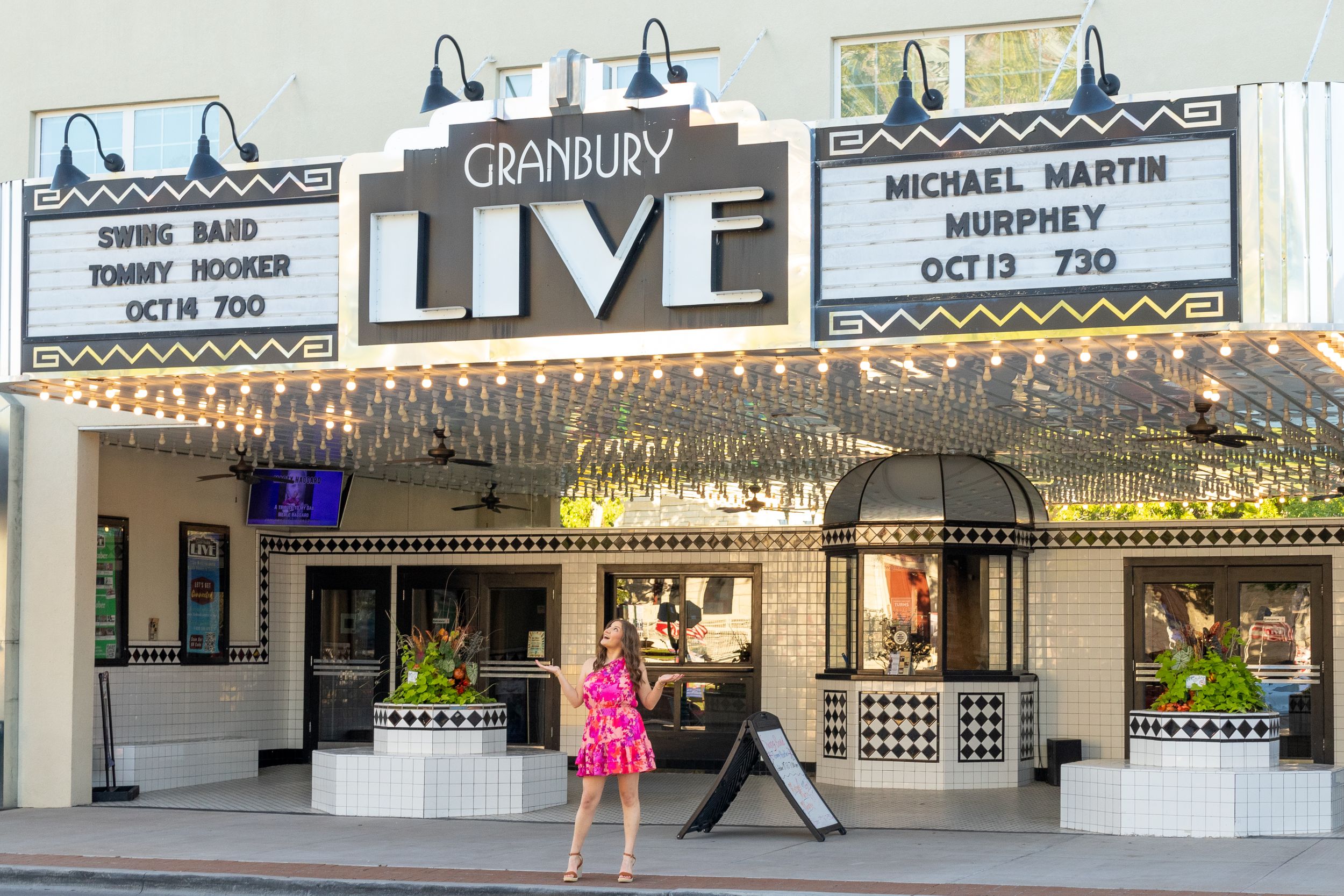 Woman in pink dress standing in front of Granbury Live theater marquee on a sunny day.