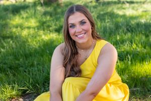 Smiling brunette woman in yellow dress sits on green grass outdoors.