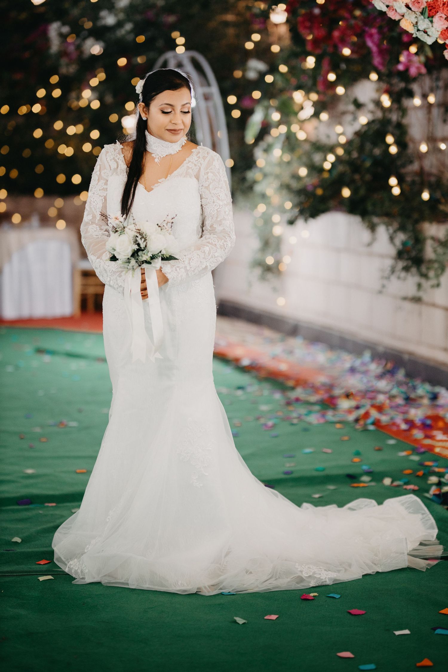 Bride in lace wedding gown poses on green carpet with scattered rose petals.