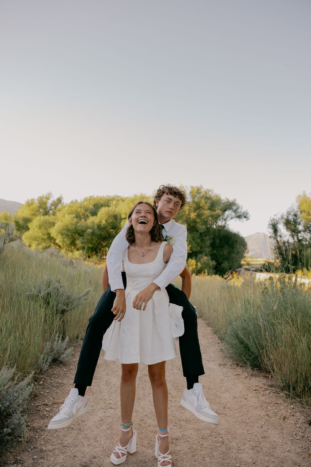 A playful couple embracing and laughing on a nature trail with trees in the background.