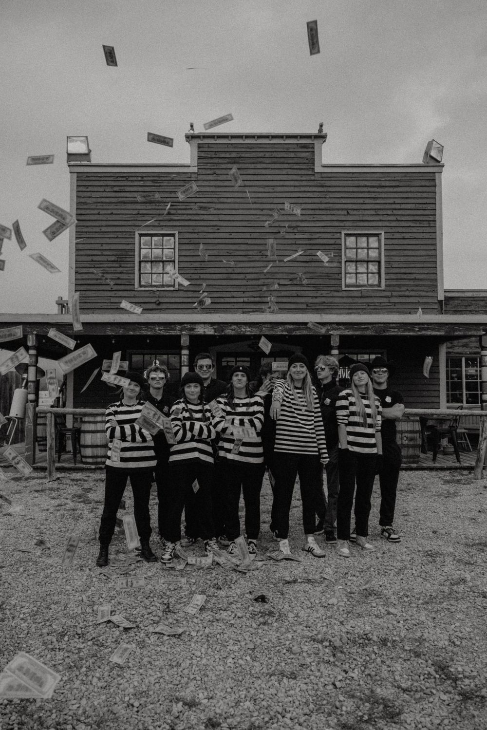 Group in prison stripes poses as money falls from sky in front of western building.