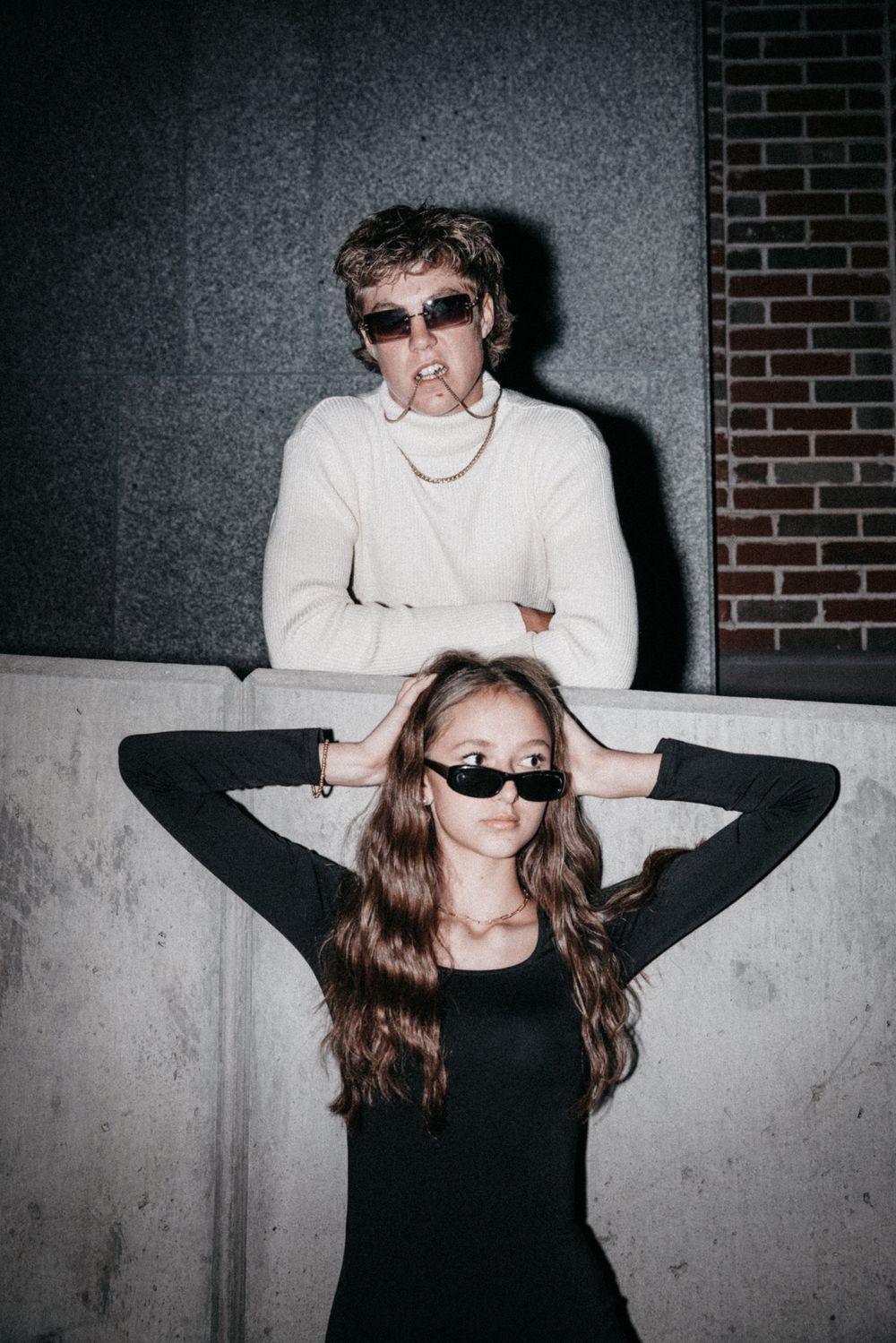 Artistic portrait of two people in black and white clothing wearing sunglasses against a concrete wall.