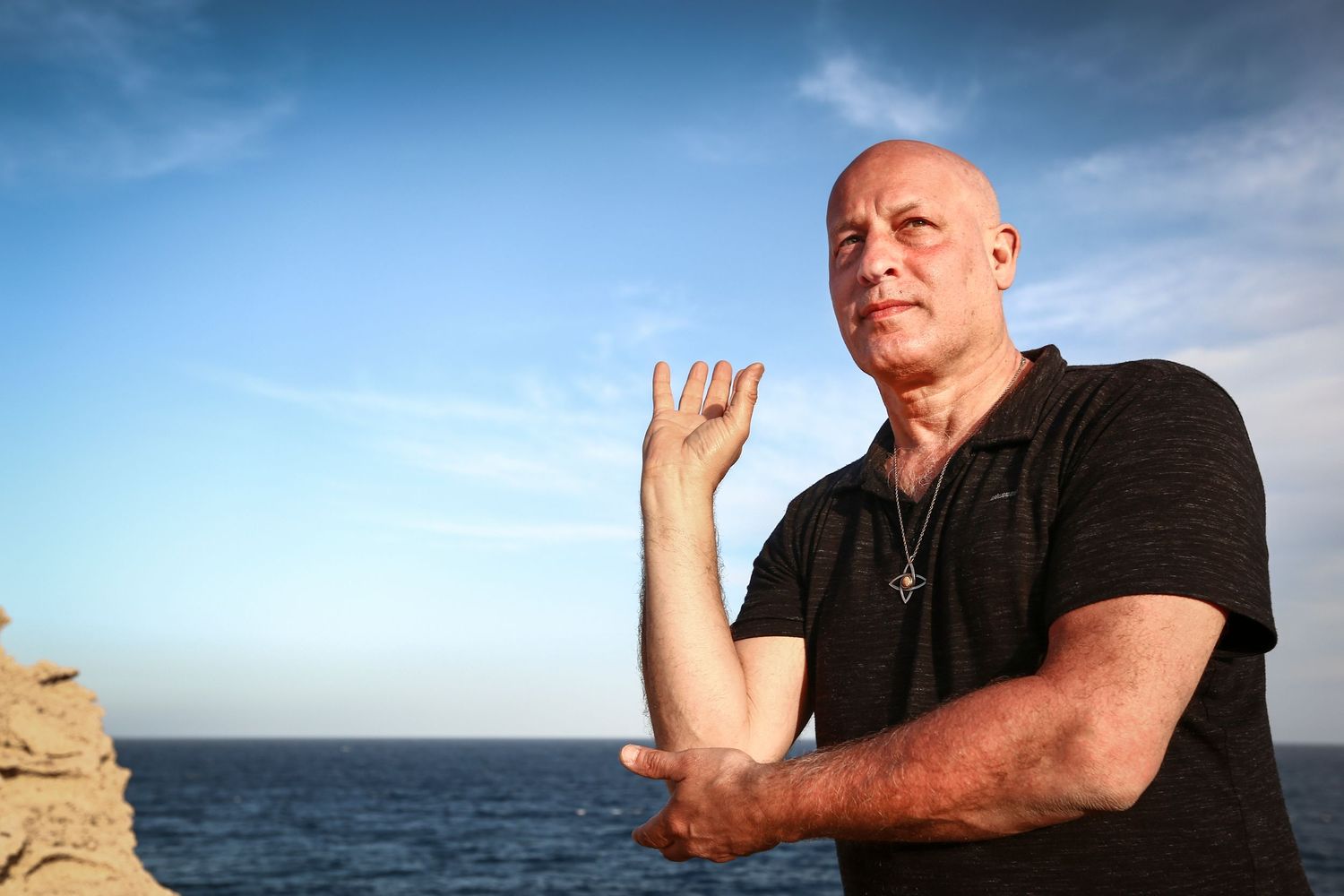 Bald man in black shirt practicing martial arts poses near the ocean under a blue sky.