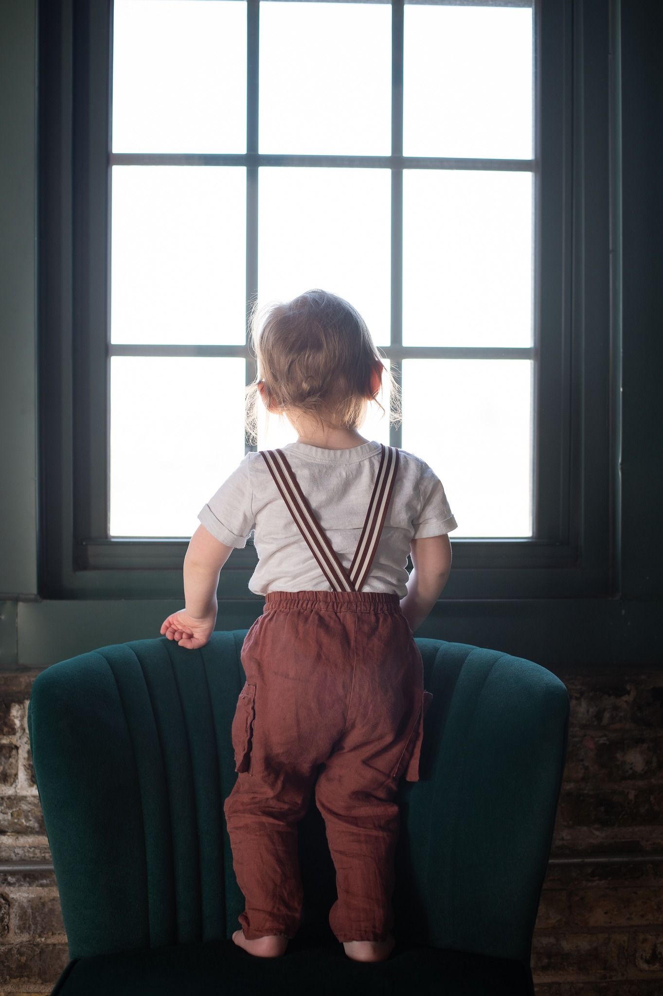 Toddler in suspender pants standing on teal chair looking out large window.