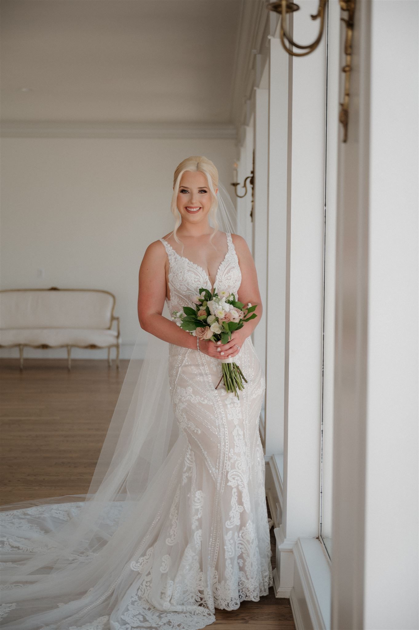 A full length portrait of a bride in a fitted lace gown holding a fresh floral bouquet.