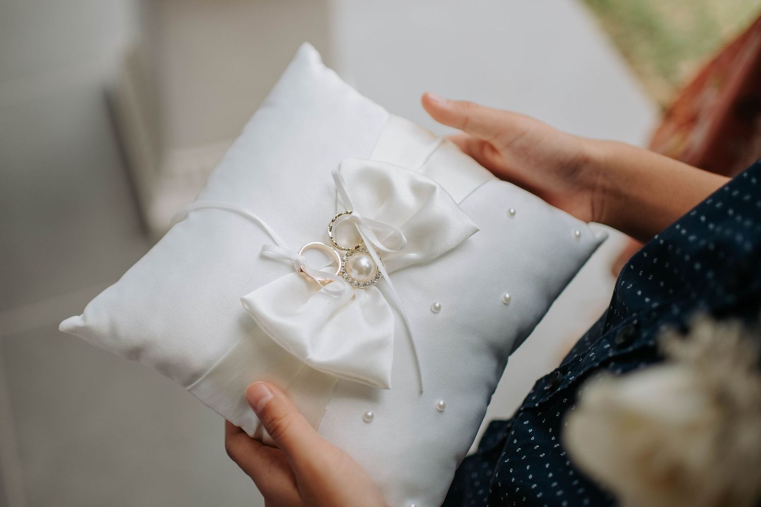 White wedding ring pillow with satin bow held against light background.