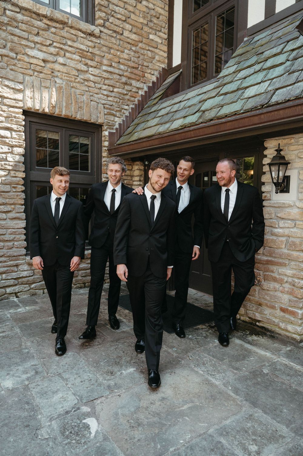 Groomsmen in black suits and ties walking confidently outside stone building entrance.