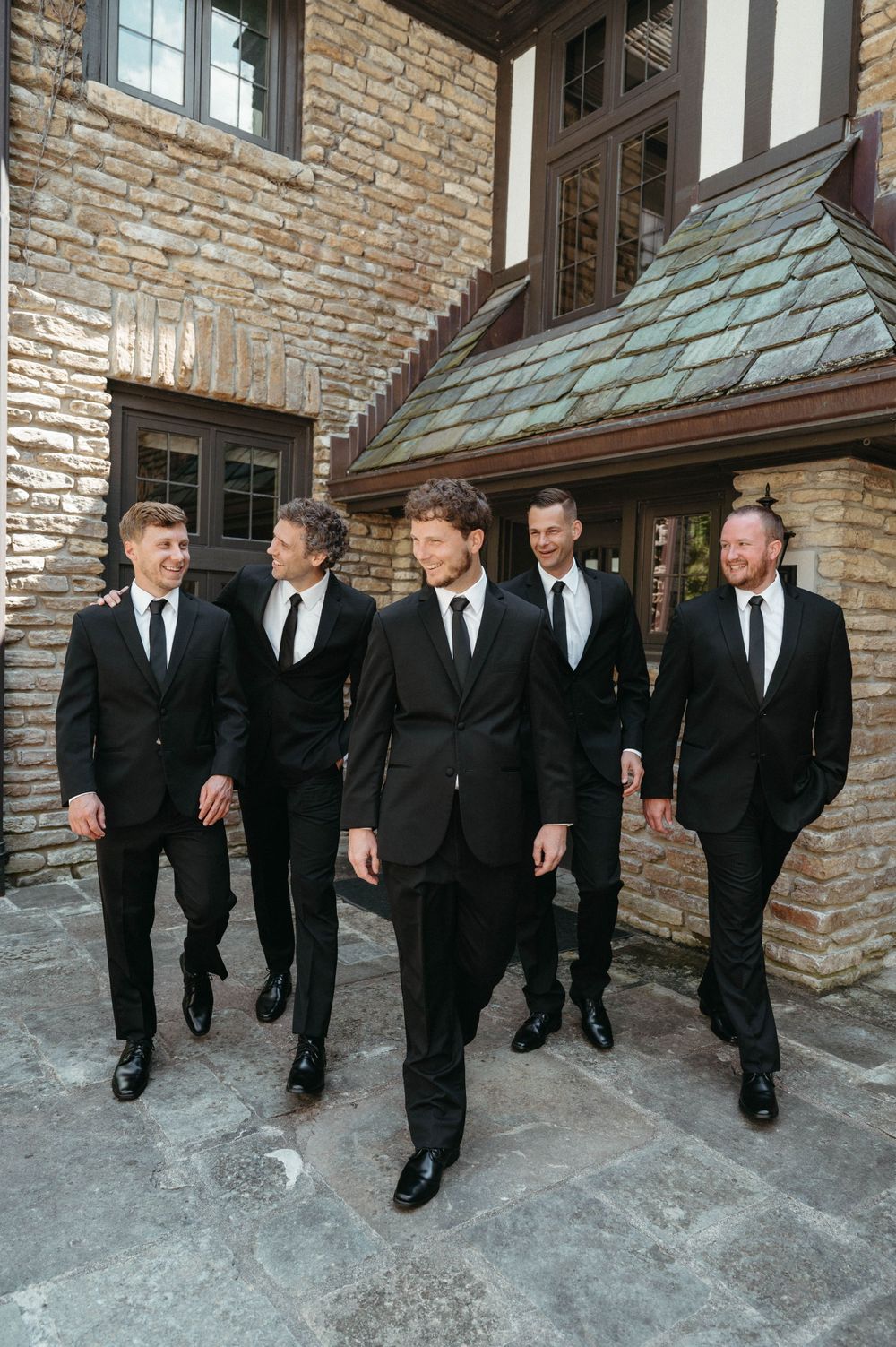 Groomsmen in matching black suits and ties walking together on stone pathway.