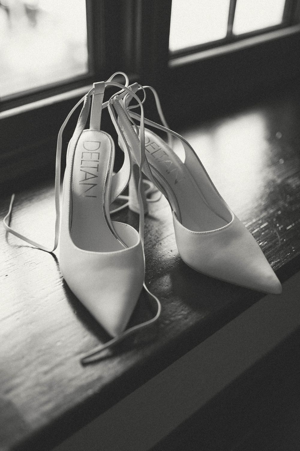 Elegant white slingback heels with pointed toes sit near a window in dramatic black and white lighting.