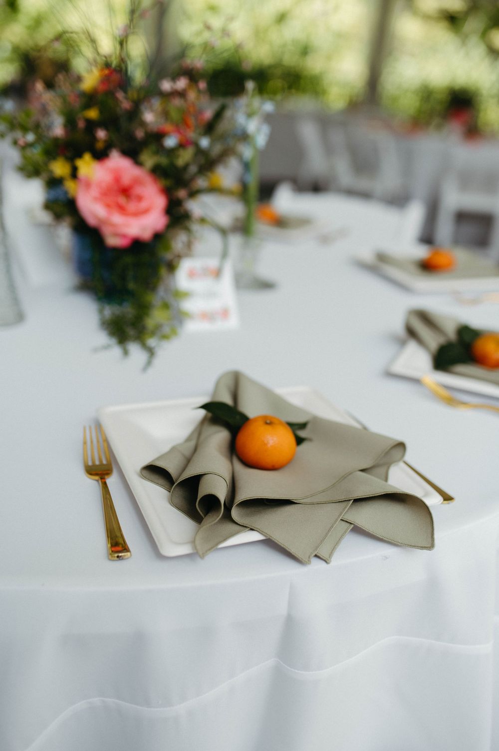 Elegant table setting with gray napkin, golden cutlery, and fresh citrus on white tablecloth with pink floral centerpiece.