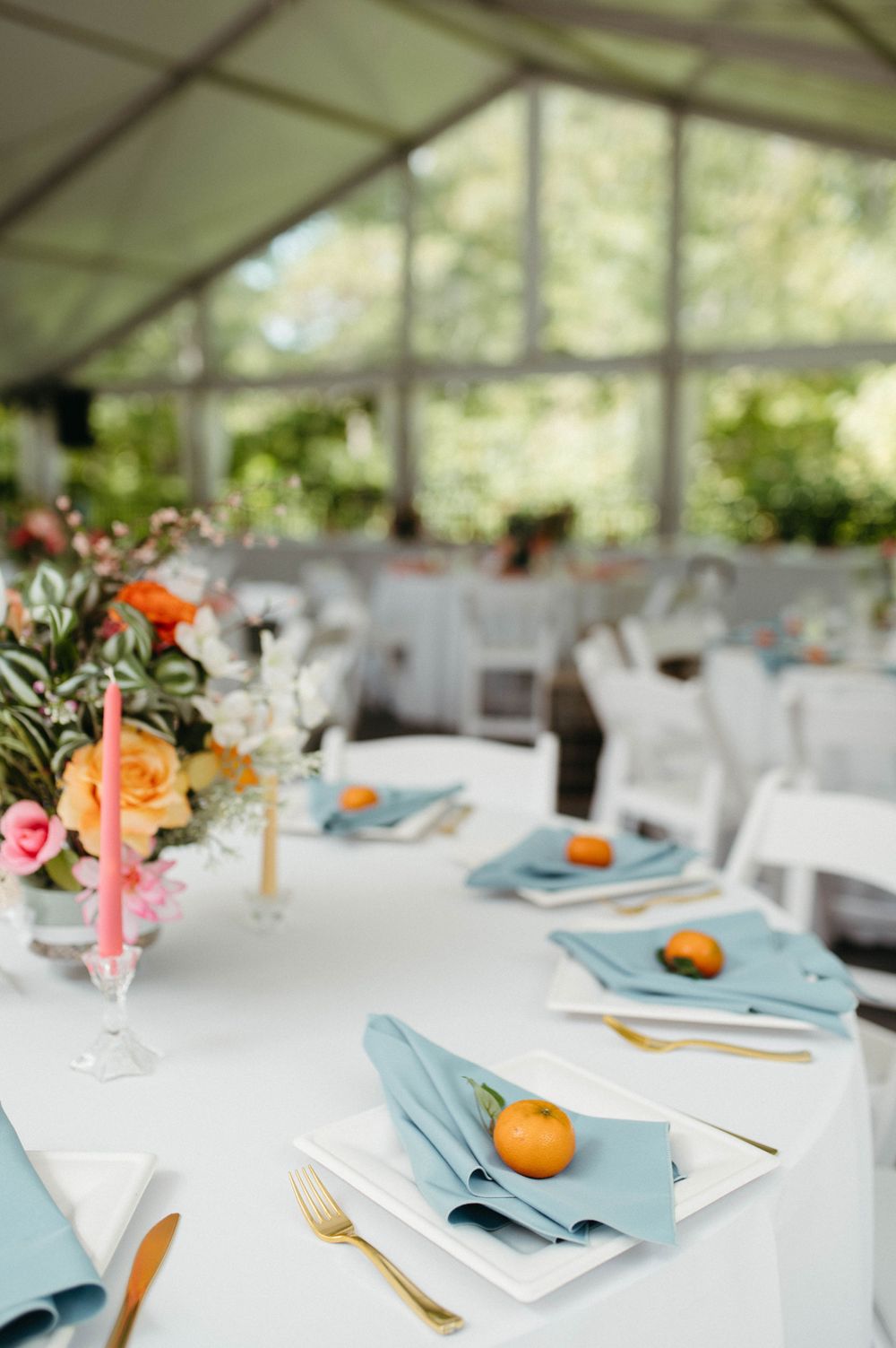Light-filled wedding reception tent with blue napkins and orange accents on white-clothed tables.